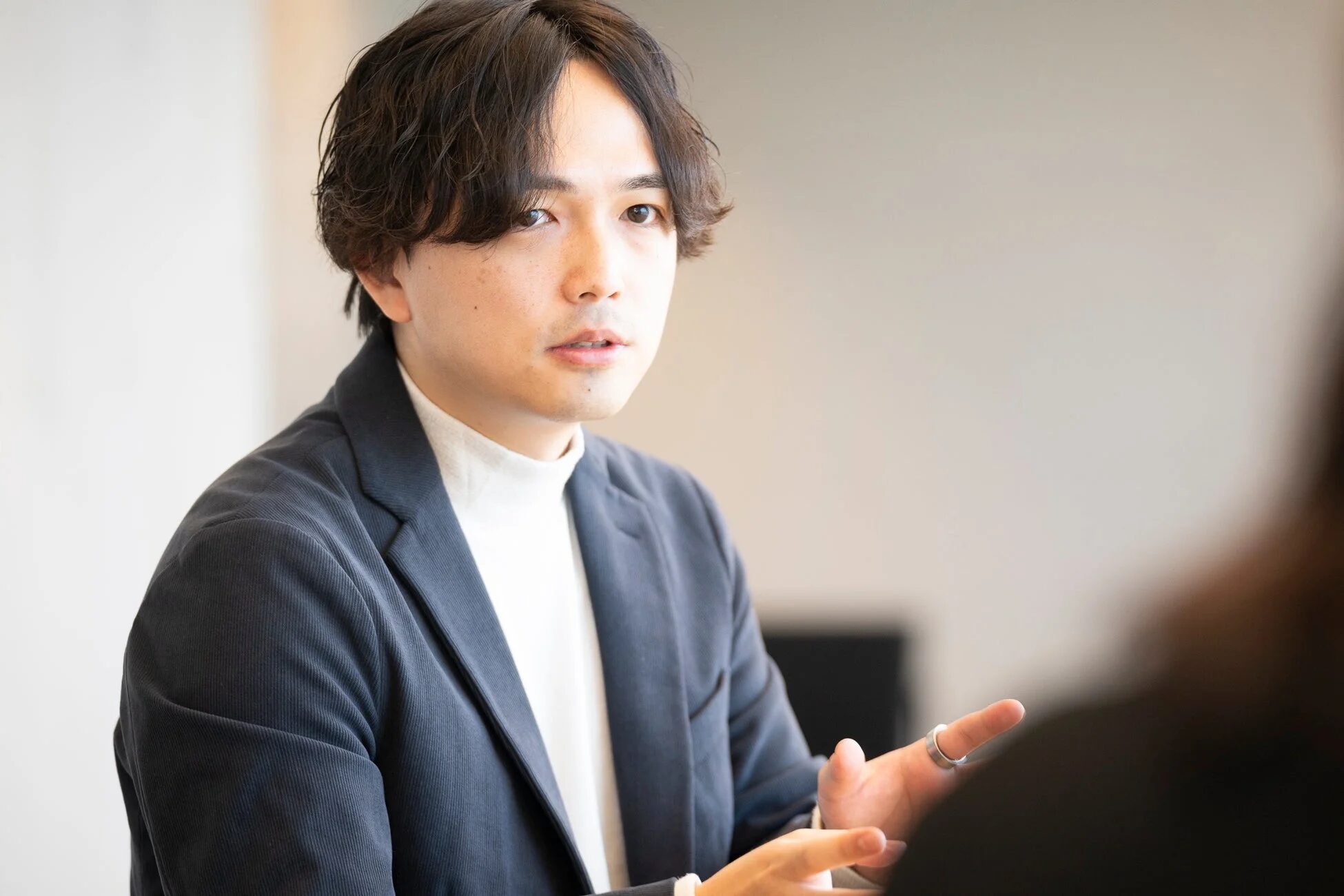 白いタートルネックと紺色のジャケットを着用し、何かを説明するように手を動かしている男性