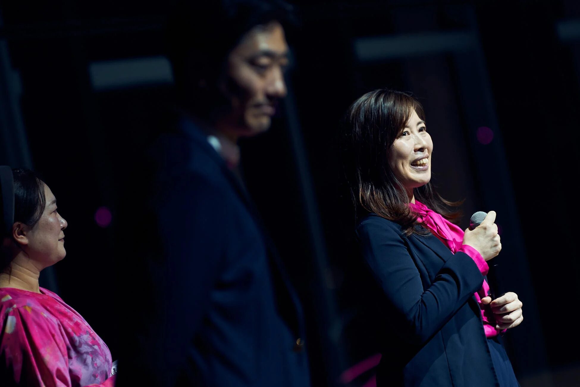暗いステージで、マイクを持ち笑顔で話すピンクのトップスを着た女性。