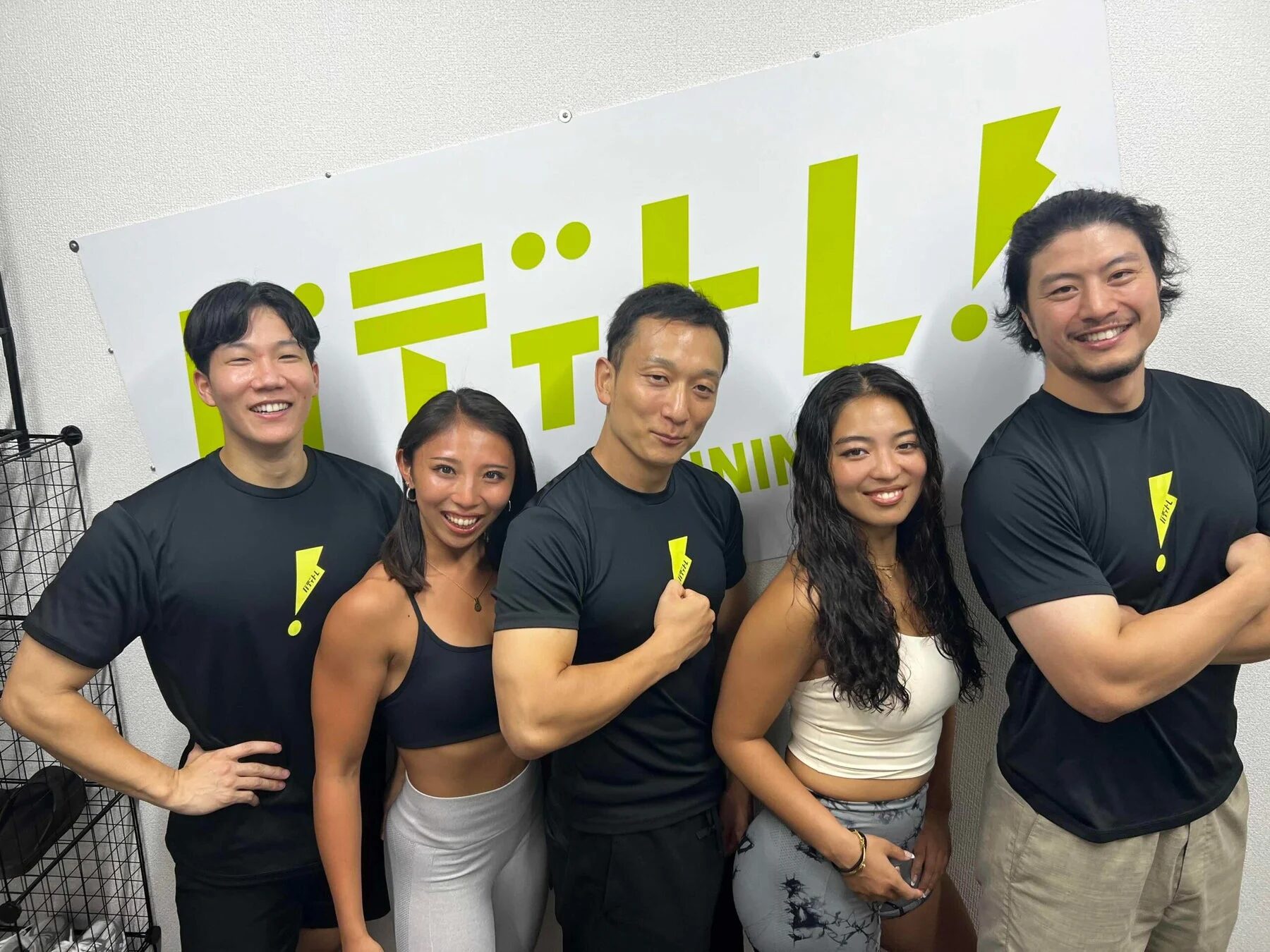 フィットネスジムのロゴを背景に、5人の男女が笑顔でポーズをとっています。彼らは全員トレーニングウェアを着用しており、健康的な雰囲気です。