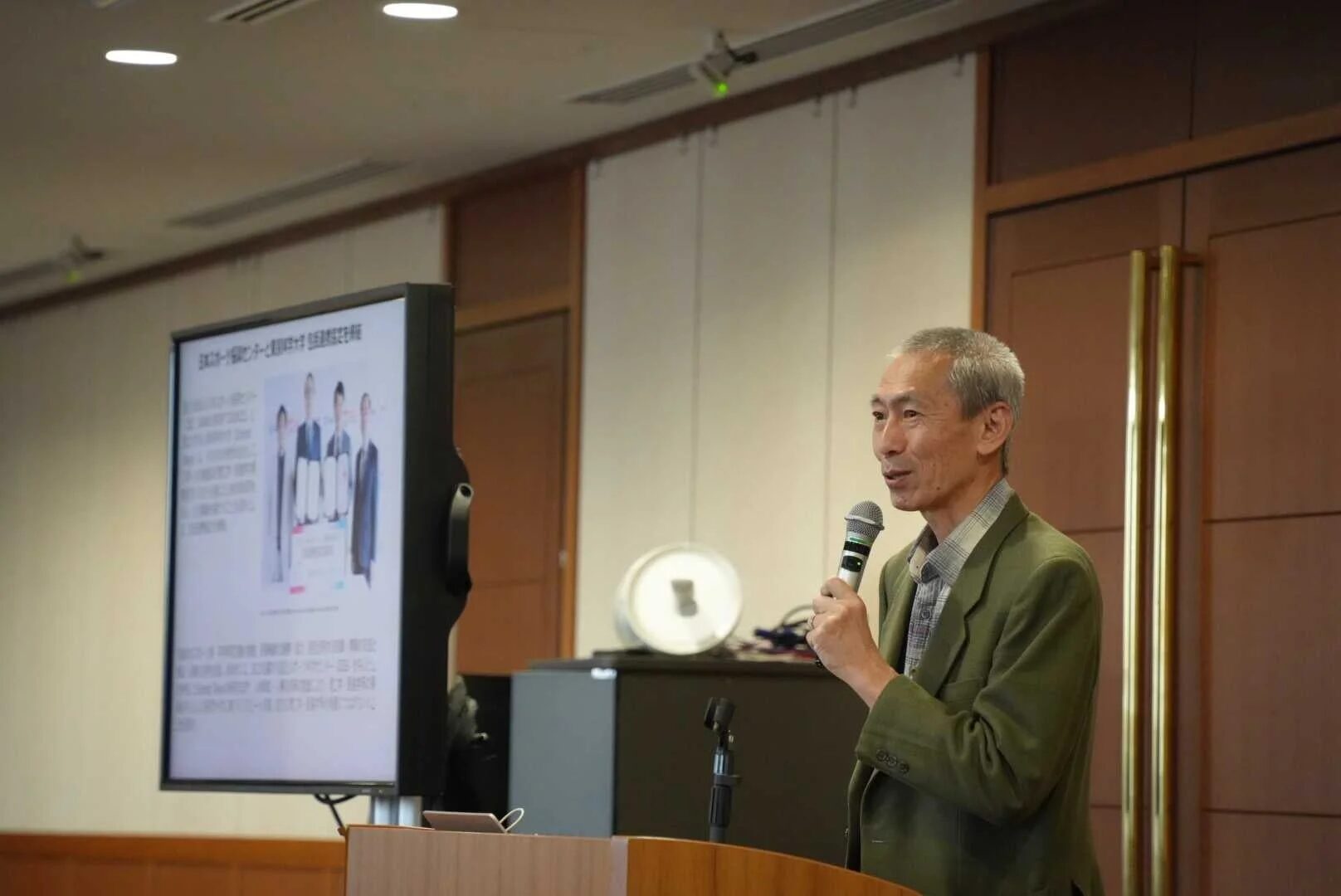 会議室のような場所で、一人の男性がマイクを持って講演している様子。