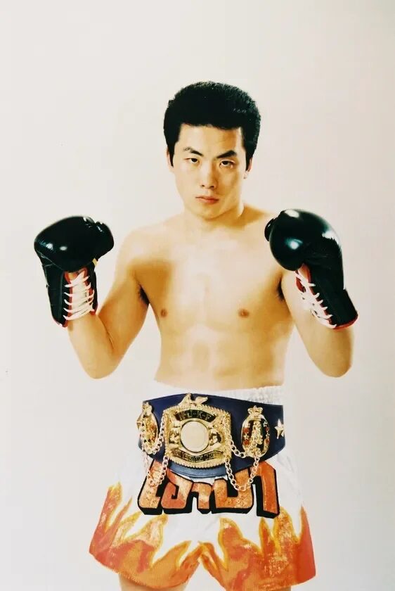 ボクシンググローブとチャンピオンベルトを身につけ、炎の模様のショートパンツを履いたアジア人男性格闘家がスタジオでポーズをとっています。