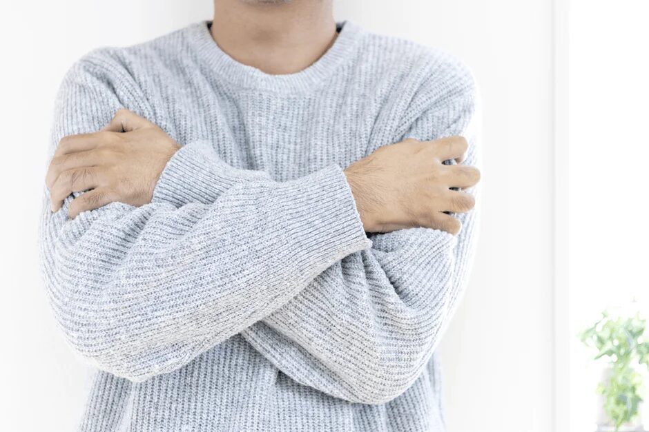 グレーのニットセーターを着た男性が腕を組み、寒そうな表情をしています。