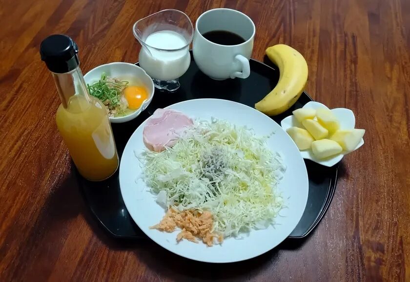 木目のテーブルに置かれた朝食のセット。千切りキャベツのサラダ、納豆と生卵、ハム、鮭フレーク、コーヒー、牛乳、バナナ、りんごなど、栄養バランスの取れた献立です。