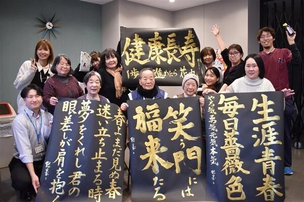 高齢者と若いスタッフが書道作品を囲んで笑顔で集合