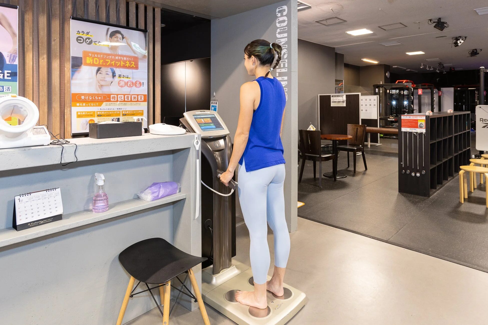 フィットネスジムで女性が体組成計を使用している様子。背景には「新Dr.フィットネス」のポスターがあり、糖化度測定などの健康診断サービスが案内されている。
