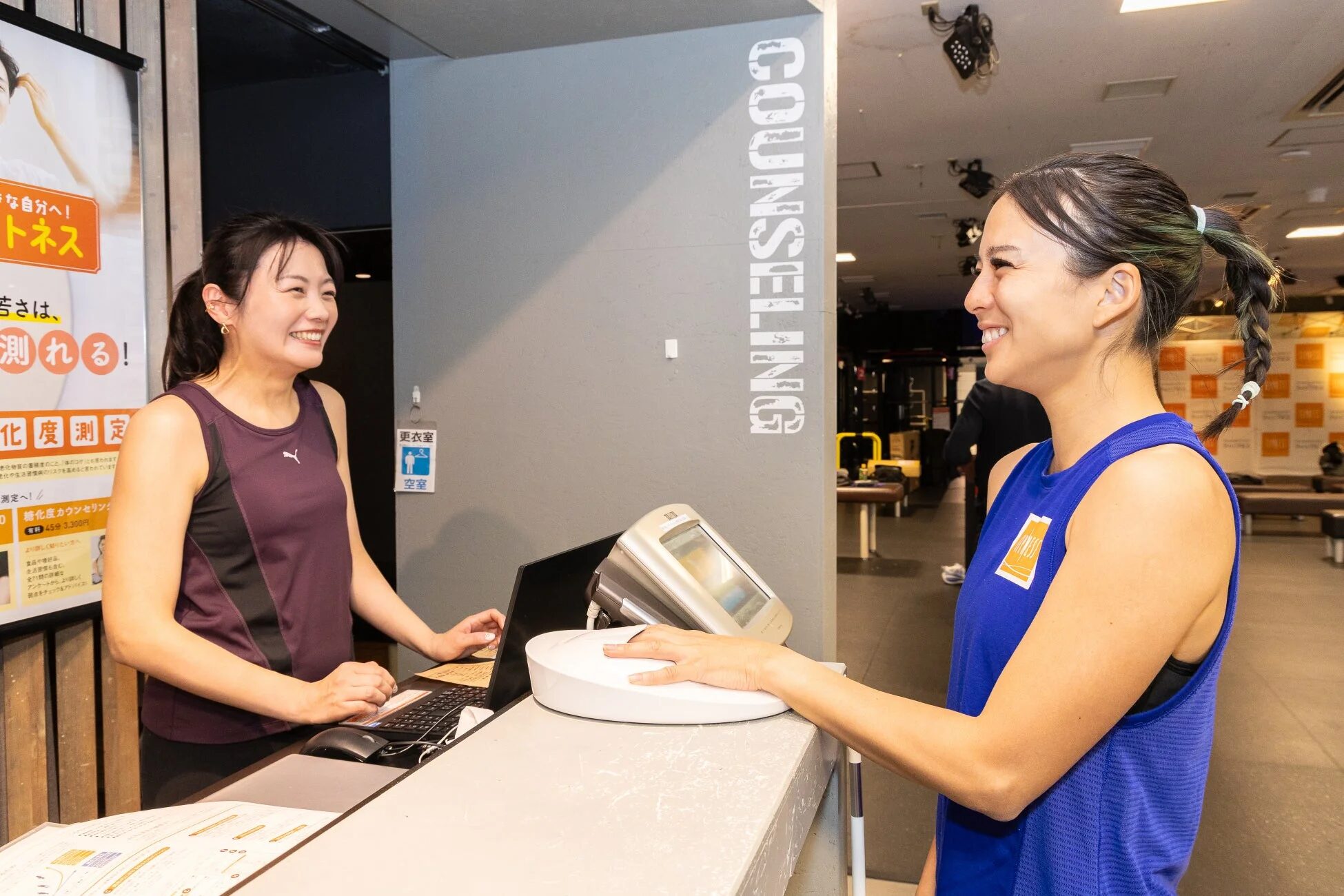 フィットネスジムの受付で、一人の女性が健康測定器に手を置き、スタッフと笑顔で会話しています。壁には「COUNSELING」の文字や「更衣室」の案内が見えます。