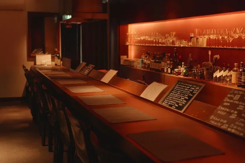 温かい照明が印象的なバーカウンターのある飲食店内部の様子。多くのグラスや酒瓶が並び、黒板にはメニューが書かれている。落ち着いた雰囲気で、夜のひとときを過ごすのに適した空間に見える。