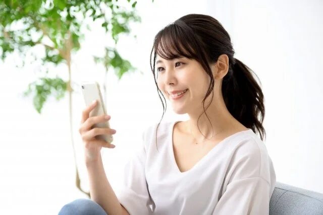 明るい室内でスマートフォンを操作しながら笑顔を見せる若い女性の姿です。リラックスした様子が伝わります。
