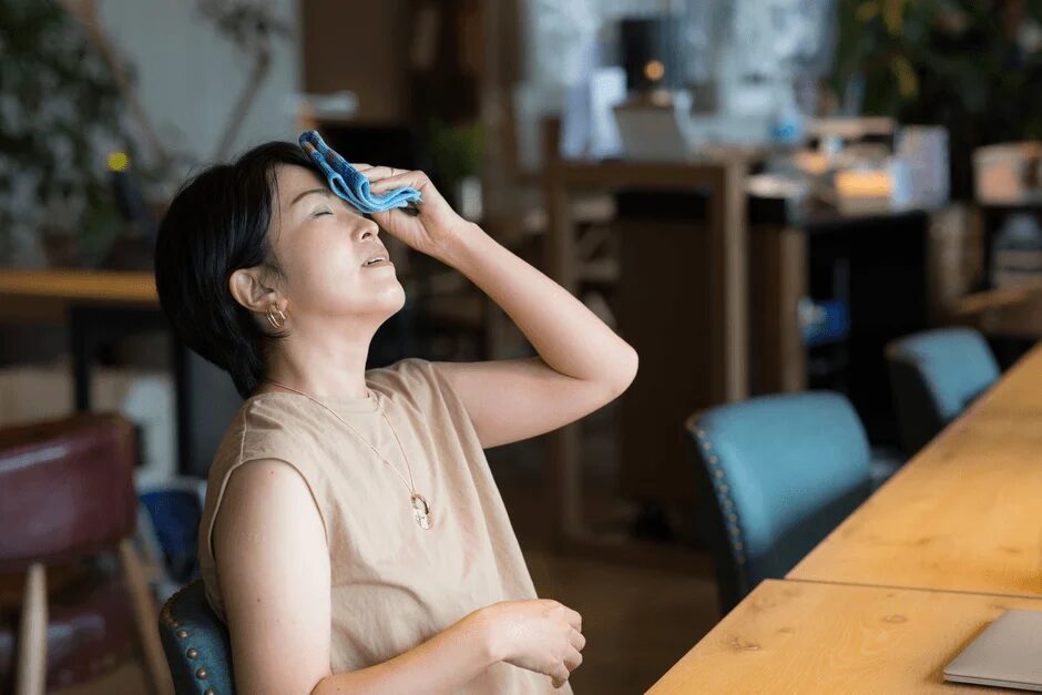 オフィスのような室内で、暑そうにしている女性が額に青いタオルを当てて汗を拭いています。熱中症対策や体調管理をしている様子がうかがえます。