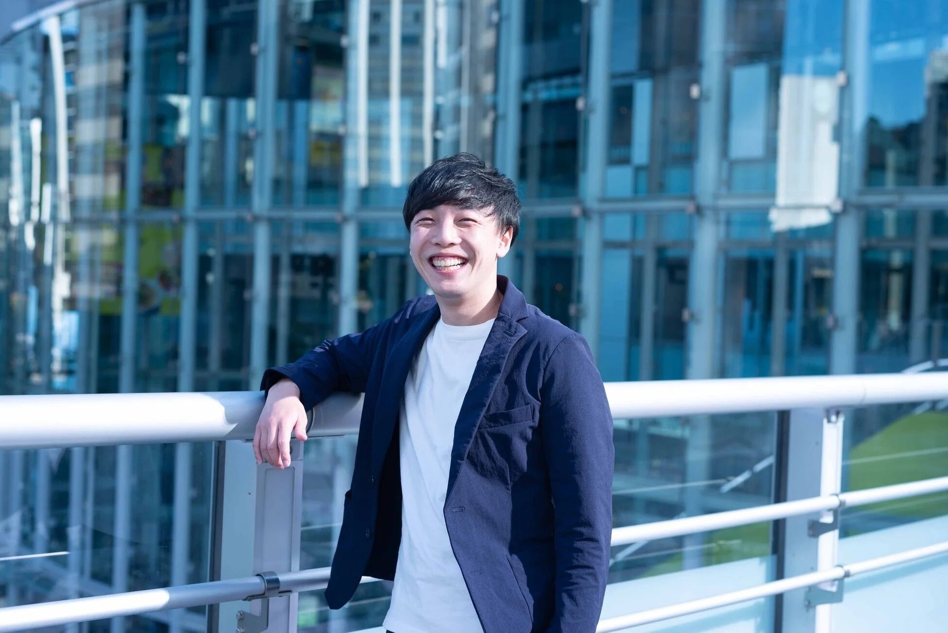 笑顔のアジア人男性が、ネイビーのジャケットと白いTシャツを着て、屋外の手すりに寄りかかっています