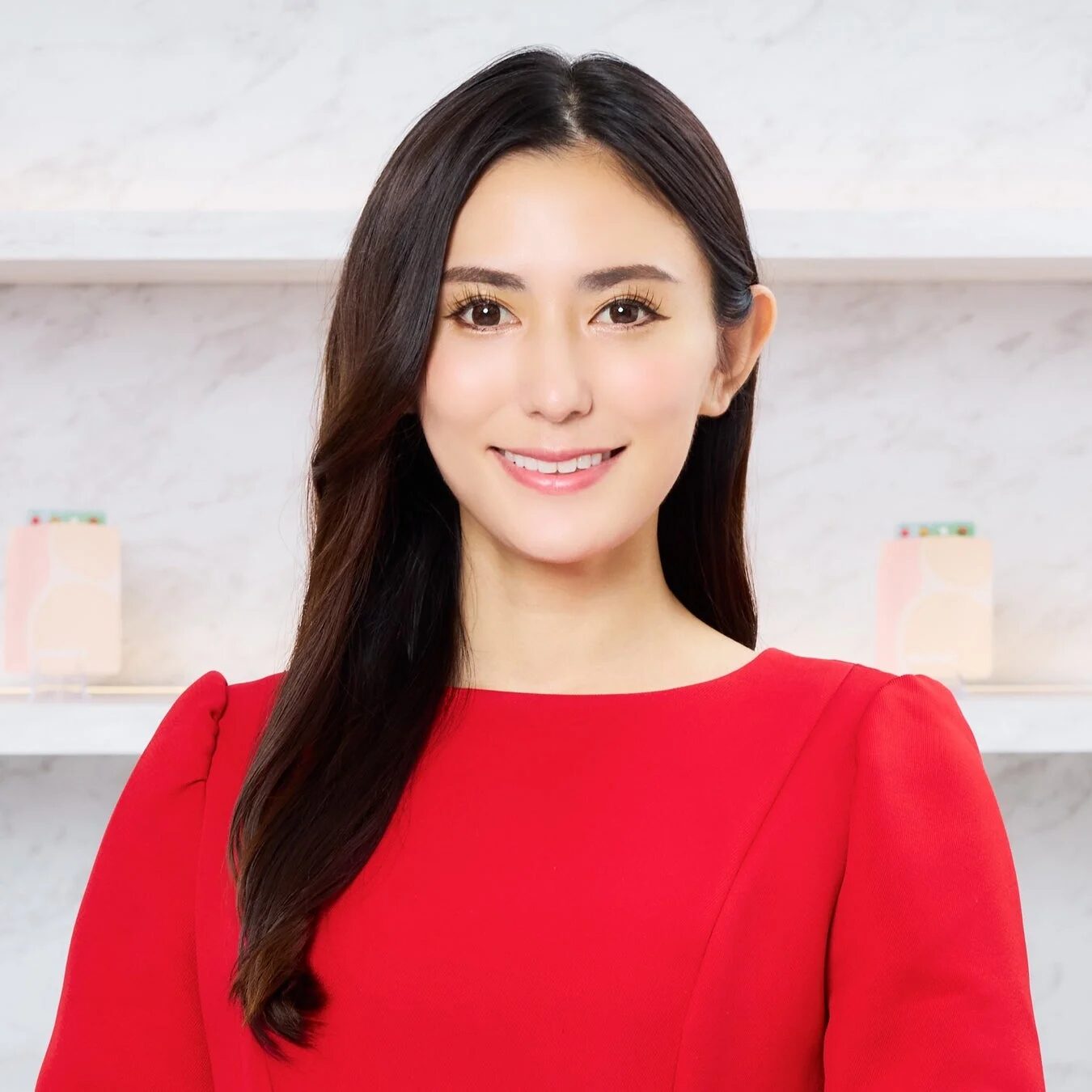 赤いドレスを着た長い黒髪の女性が、白を基調とした背景の前で笑顔を見せています。