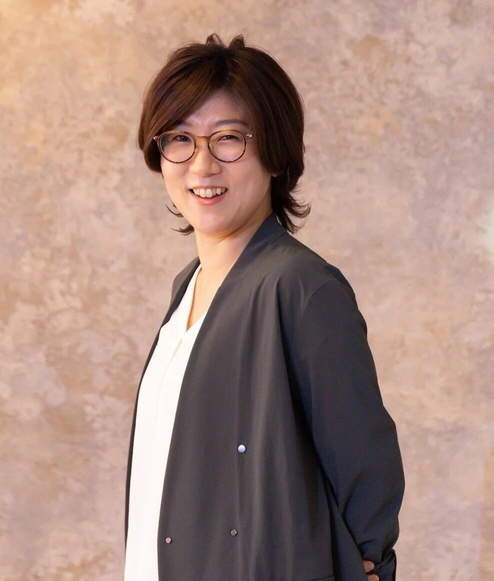 茶色の背景を背に、眼鏡をかけたショートヘアの女性が笑顔でカメラを見ているポートレート