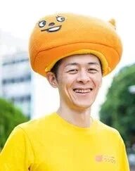 大きなオレンジ色のキャラクター帽子をかぶり、黄色いTシャツを着た男性が笑顔で写っています