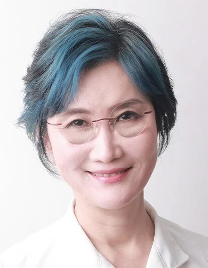 青いメッシュのショートヘアとリムレスメガネをかけたアジア人女性のポートレートです。白いシャツを着用し、明るく微笑んでいます。