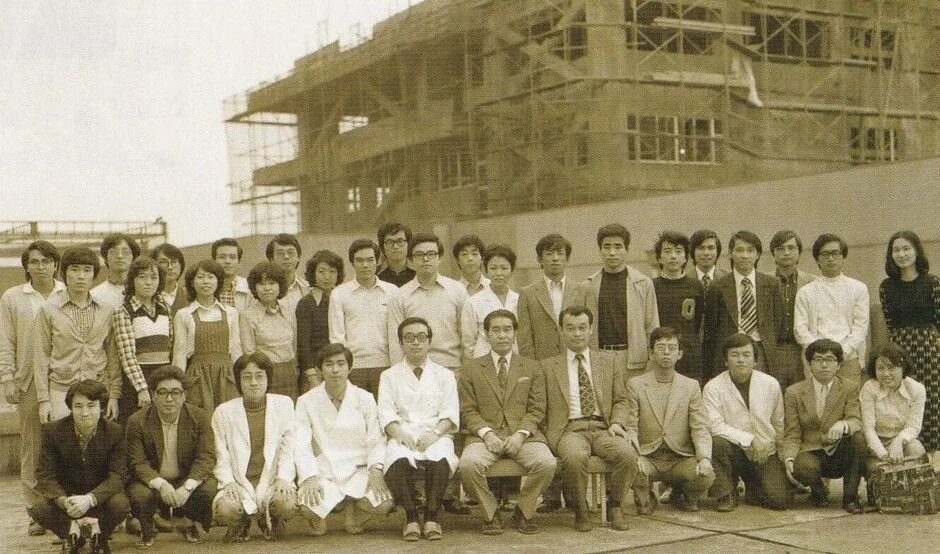 建設中の建物を背景に、多くの日本人男性と数人の女性が集合写真を撮っています。服装や雰囲気から1970年代から1980年代初頭の日本の大学や企業グループの様子を捉えたものと推測されます。