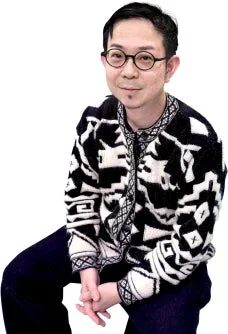 眼鏡をかけた男性が、白と黒の幾何学模様のカーディガンを着て座っています