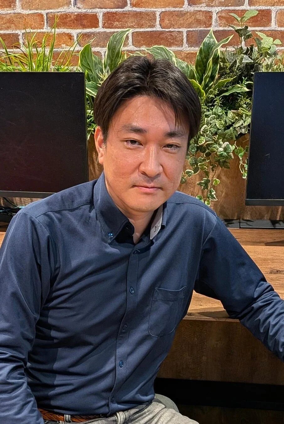紺色のシャツを着たアジア人男性が、レンガの壁と観葉植物を背景に座っているポートレート写真