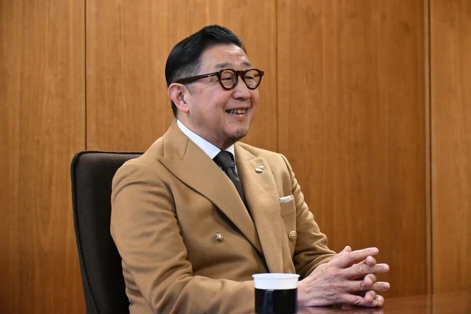 眼鏡をかけた男性が、キャメル色のジャケットを着てテーブルに座り、笑顔を見せています。木製の壁を背景に、リラックスした雰囲気で会話しているようです。