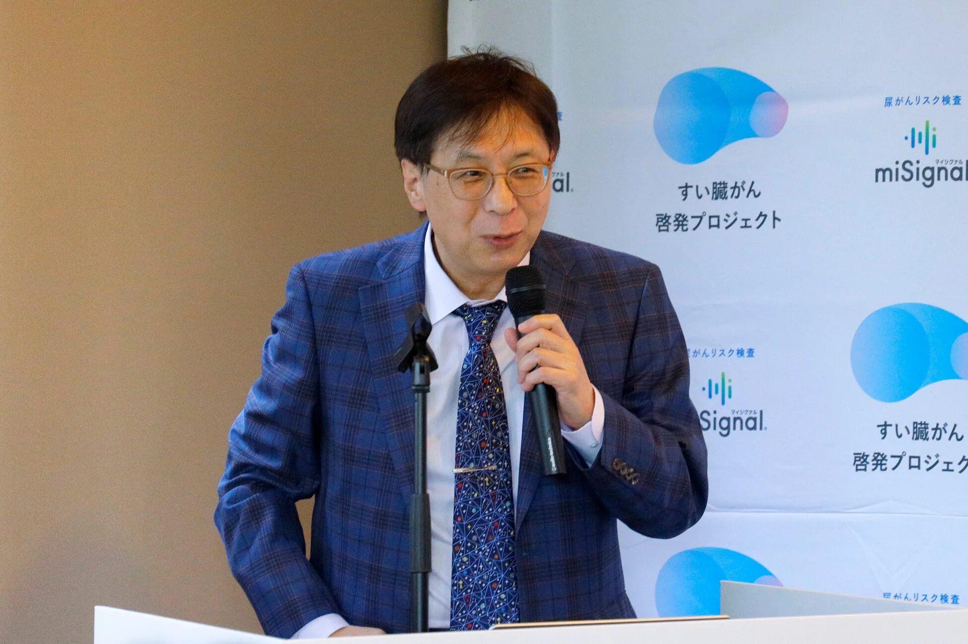 青いチェックのスーツを着た男性がマイクを持って話している様子です。背景には「すい臓がん啓発プロジェクト」と「miSignal 尿がんリスク検査」の文字が見え、医療関連のイベントでの講演風景と推測されます。