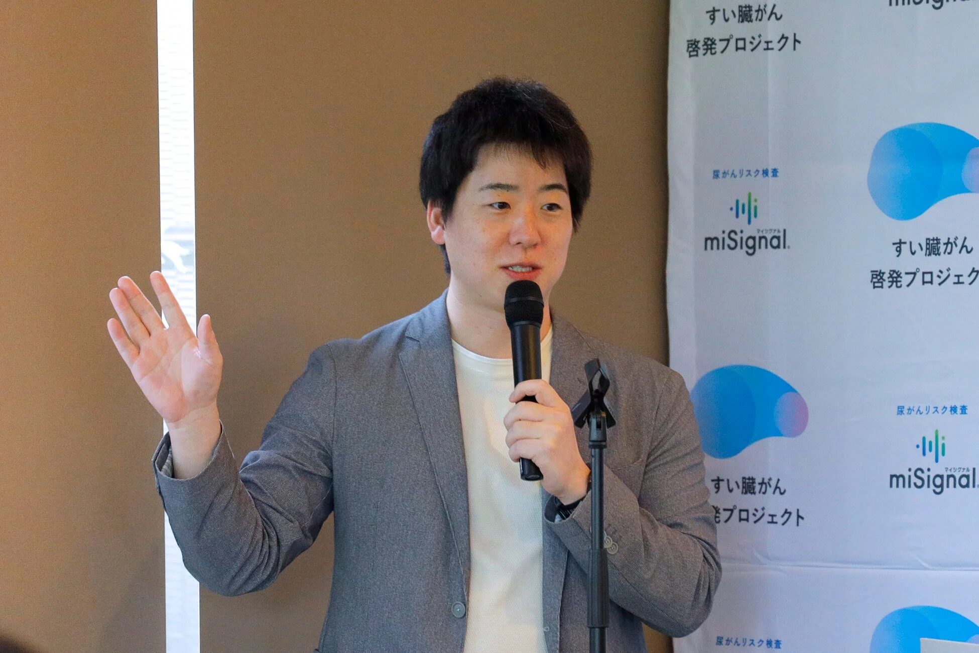 マイクを手にスピーチをする若い男性の姿。背景には「すい臓がん啓発プロジェクト」や「尿がんリスク検査 miSignal」と書かれたバナーがあり、医療関連のイベントでの登壇風景と考えられる。